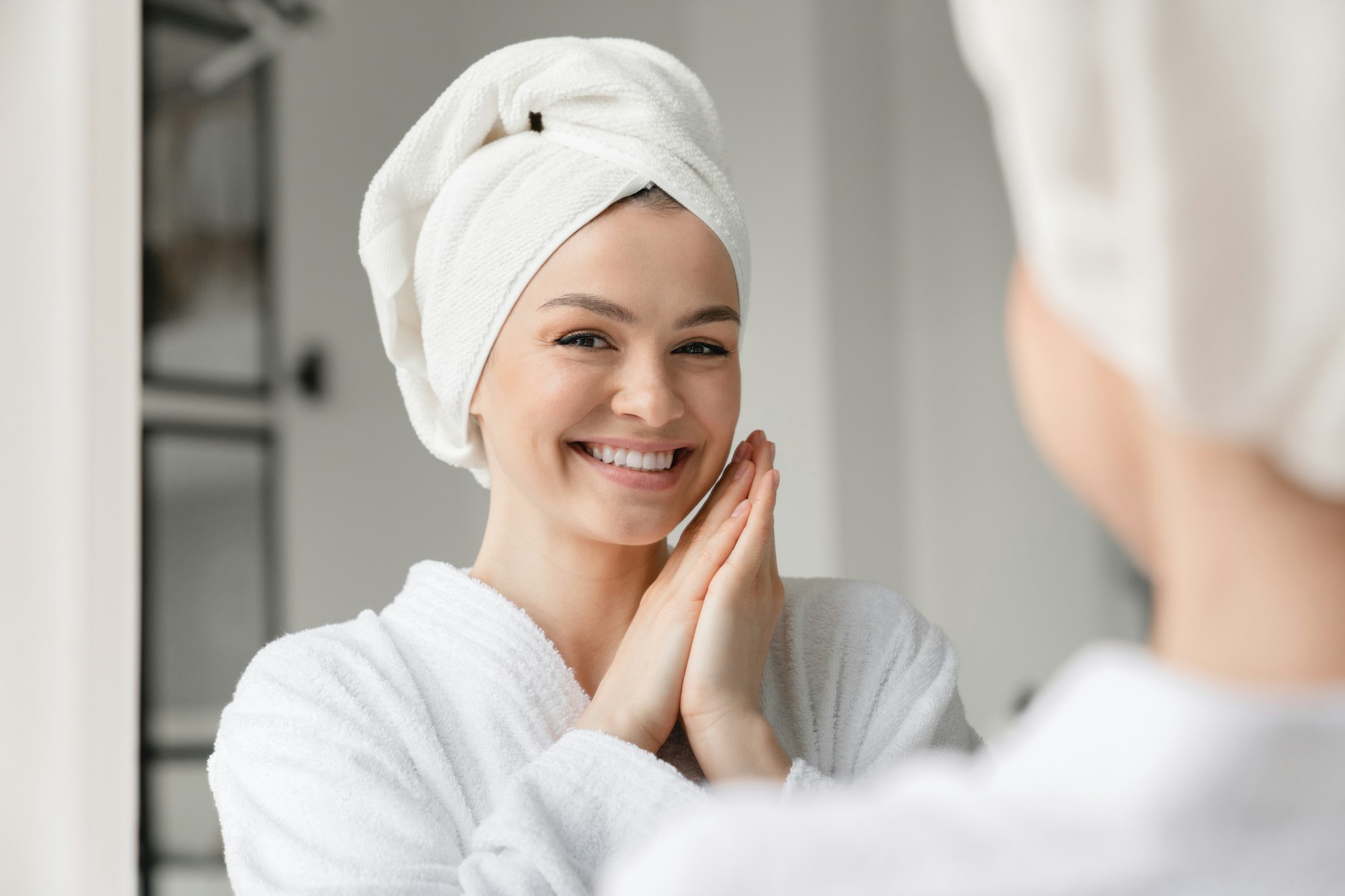 Happy white woman with toothy smile clean young clear skin in spa bath towel looking at the mirror.